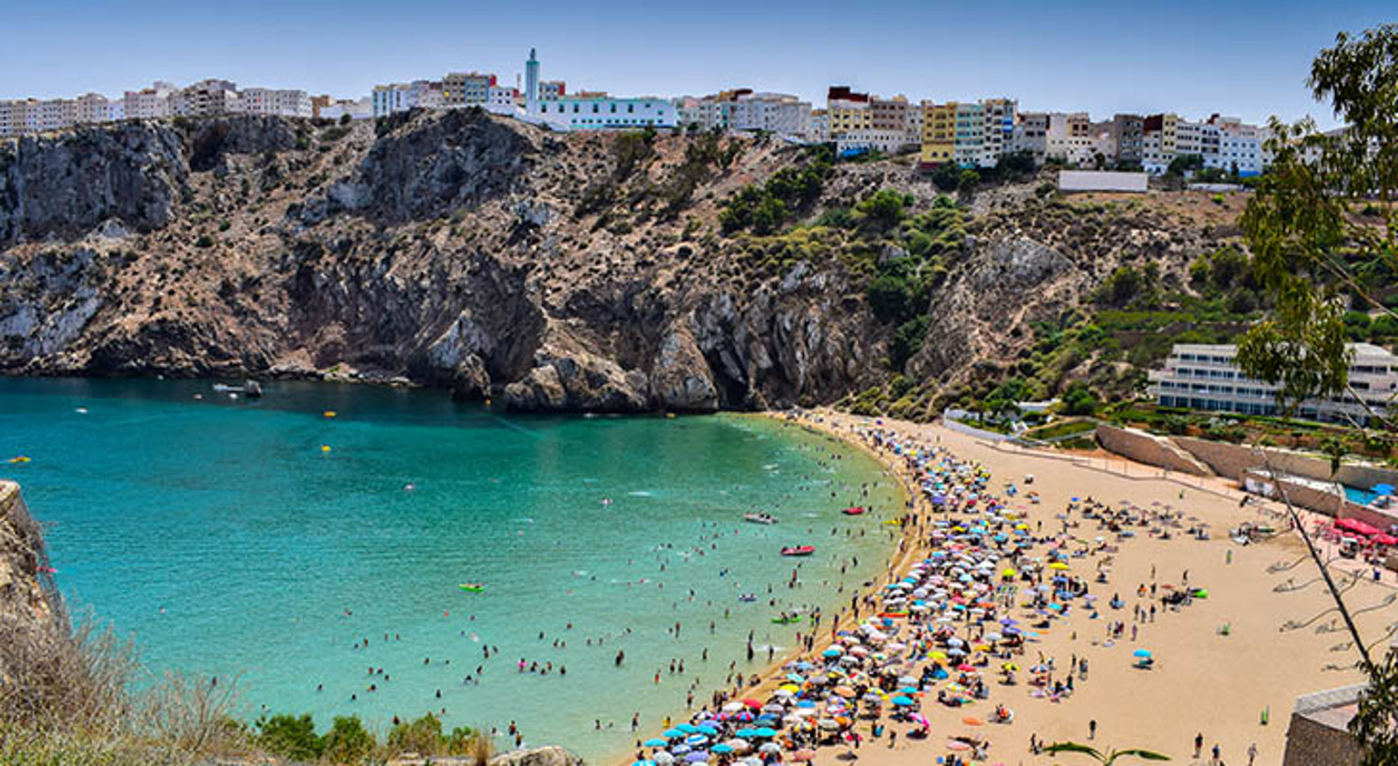 Al Hoceima Druk strand in Al Hoceima, Marokko, met turquoise baai, rotsachtige kliffen en stad op de heuvel.