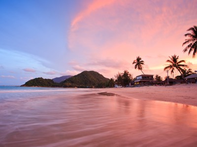 Zonsondergang op een strand in Zuid-Thailand met palmbomen, zee en heuvels op de achtergrond.