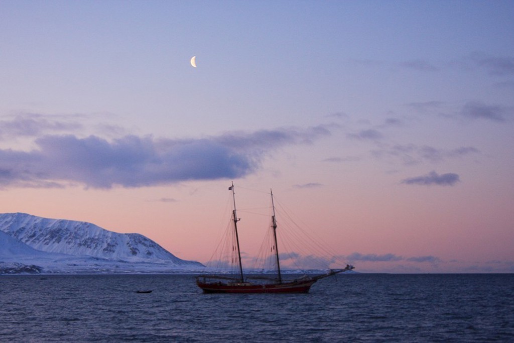 Zeilschip Noorderlicht bij Spitsbergen, met besneeuwde bergen en halve maan bij zonsondergang.