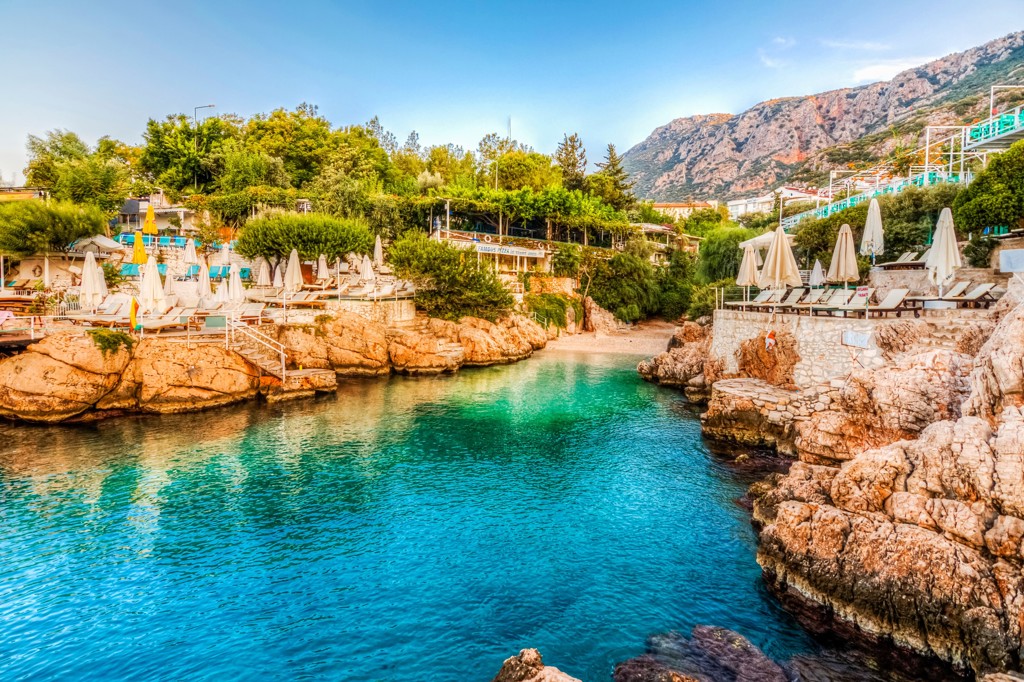 Turquoise baai met rotsen, strandbedden en parasols aan de kust van Turkije, met bergen op de achtergrond.