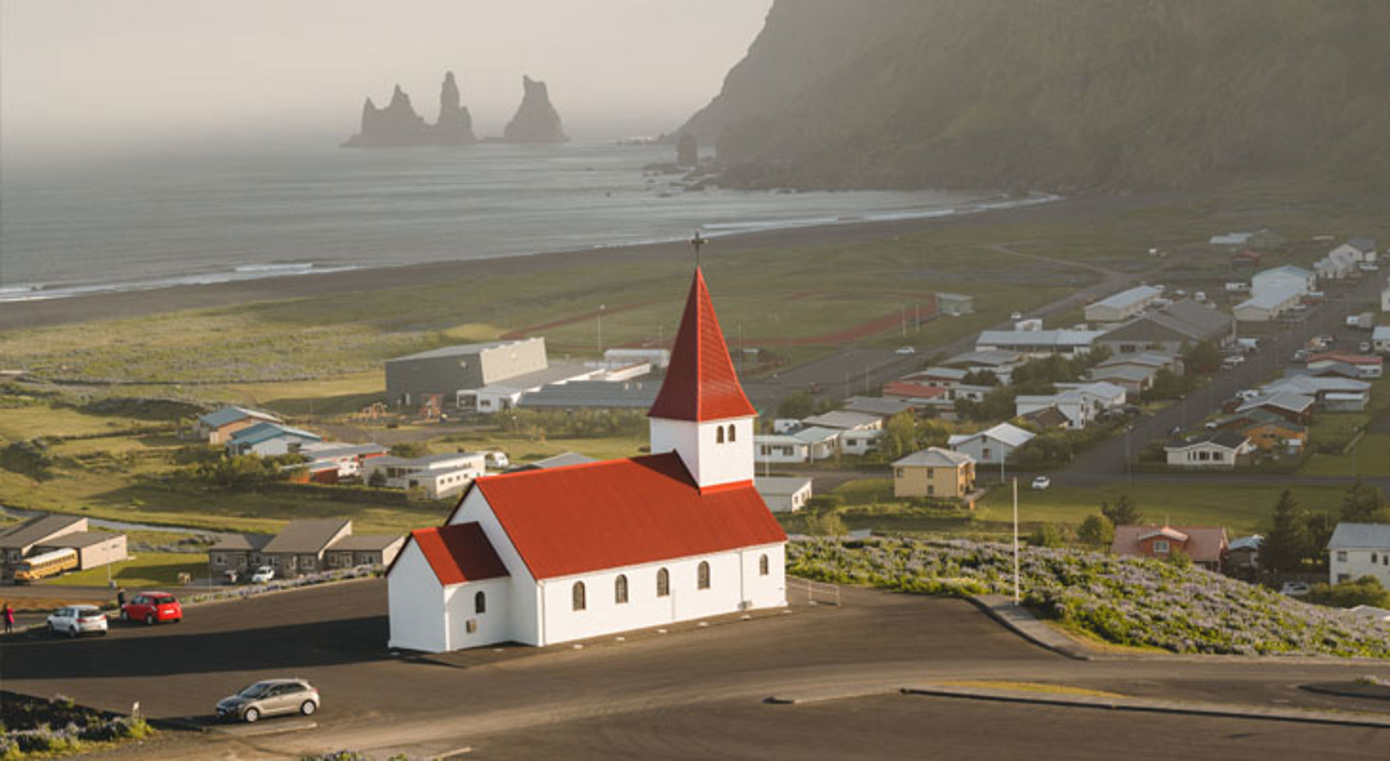 Vik IJsland Witte kerk met rood dak in Vík í Mýrdal, met zwart zandstrand en Reynisdrangar rotsen op de achtergrond.