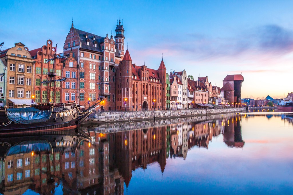 Historische kade in Gdansk met kleurrijke gevels en de Kraanpoort, weerspiegeld in het water bij zonsondergang.