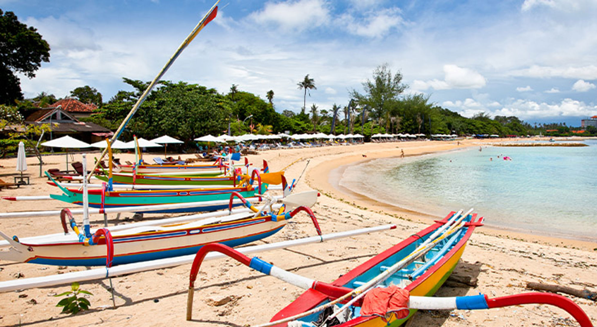 Sanur Beach Bali Kleurrijke vissersboten op het zandstrand bij een rustige baai met parasols en palmbomen.