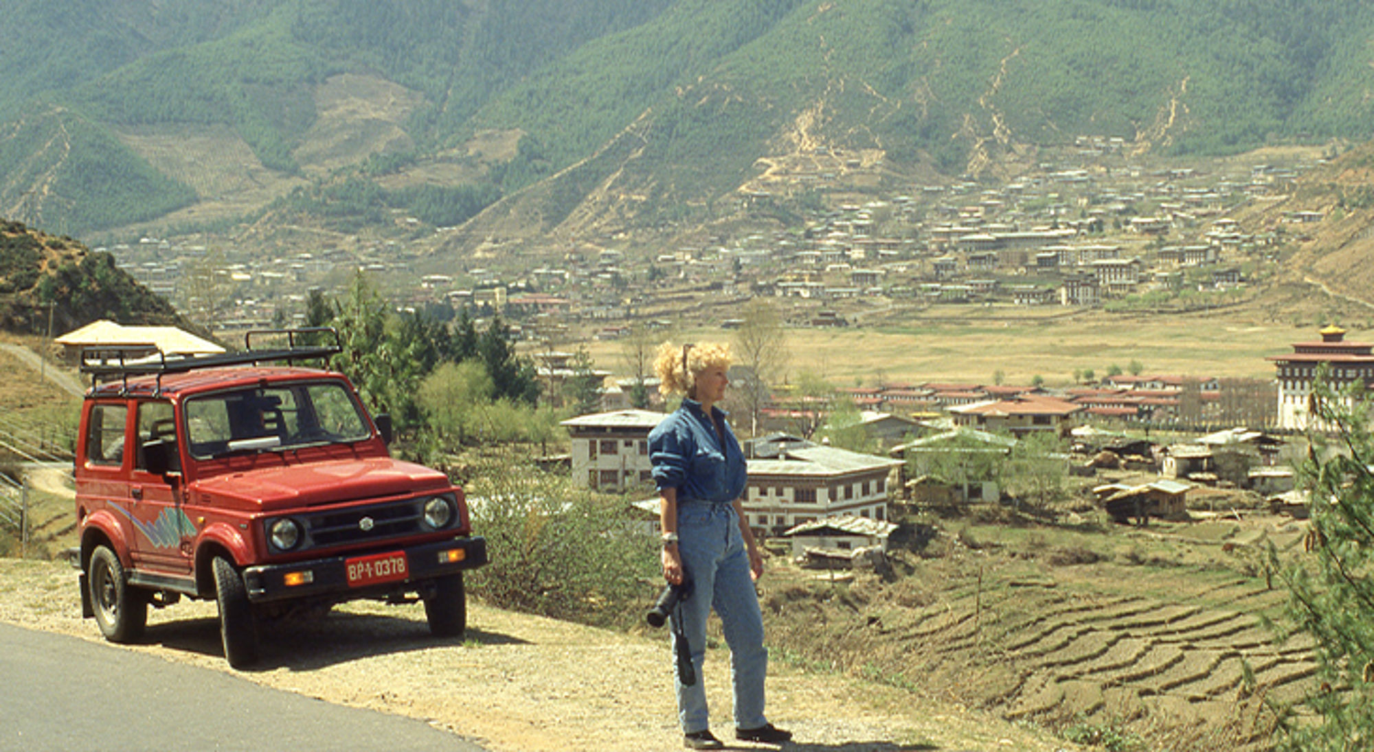Mieke in Bhutan Reiziger met camera naast rode 4x4 met uitzicht op een vallei en dorp in de bergen.