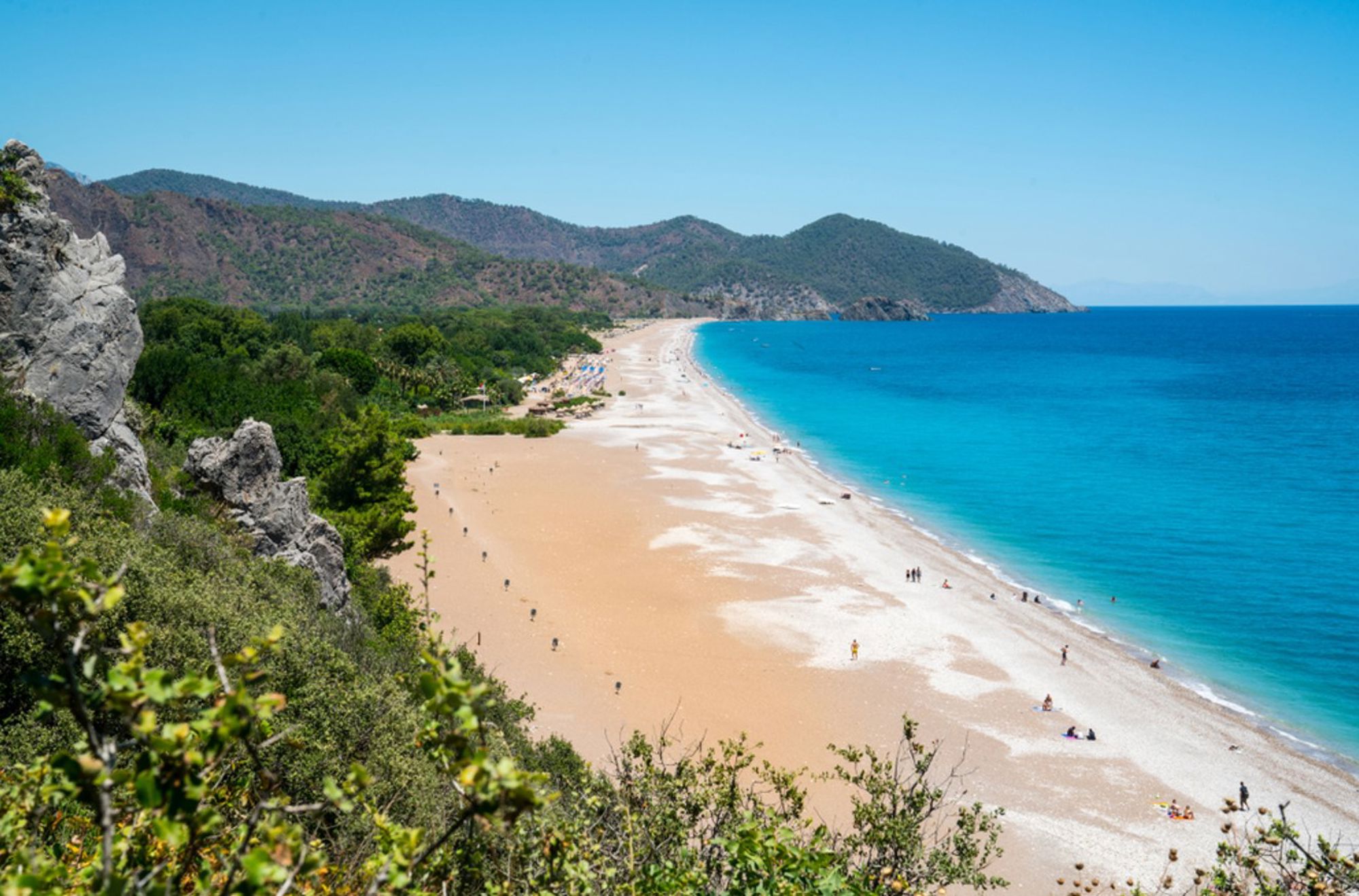 Panoramisch uitzicht op een lang zandstrand met helderblauwe zee en groene bergen aan de Turkse kust.
