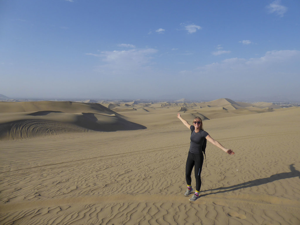 Vrouw op reis in Peru met armen wijd, staand in uitgestrekte zandduinen onder een blauwe lucht.