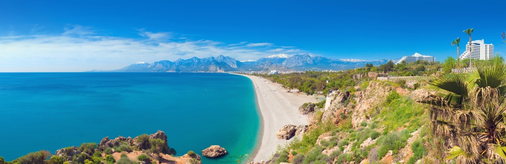 Panorama van de turquoise kust in Turkije met lang zandstrand, helderblauwe zee en bergen op de achtergrond.