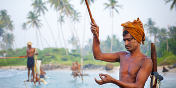 Paalvisser in Sri Lanka werpt zijn lijn uit vanaf een paal in zee, met palmbomen op de achtergrond.