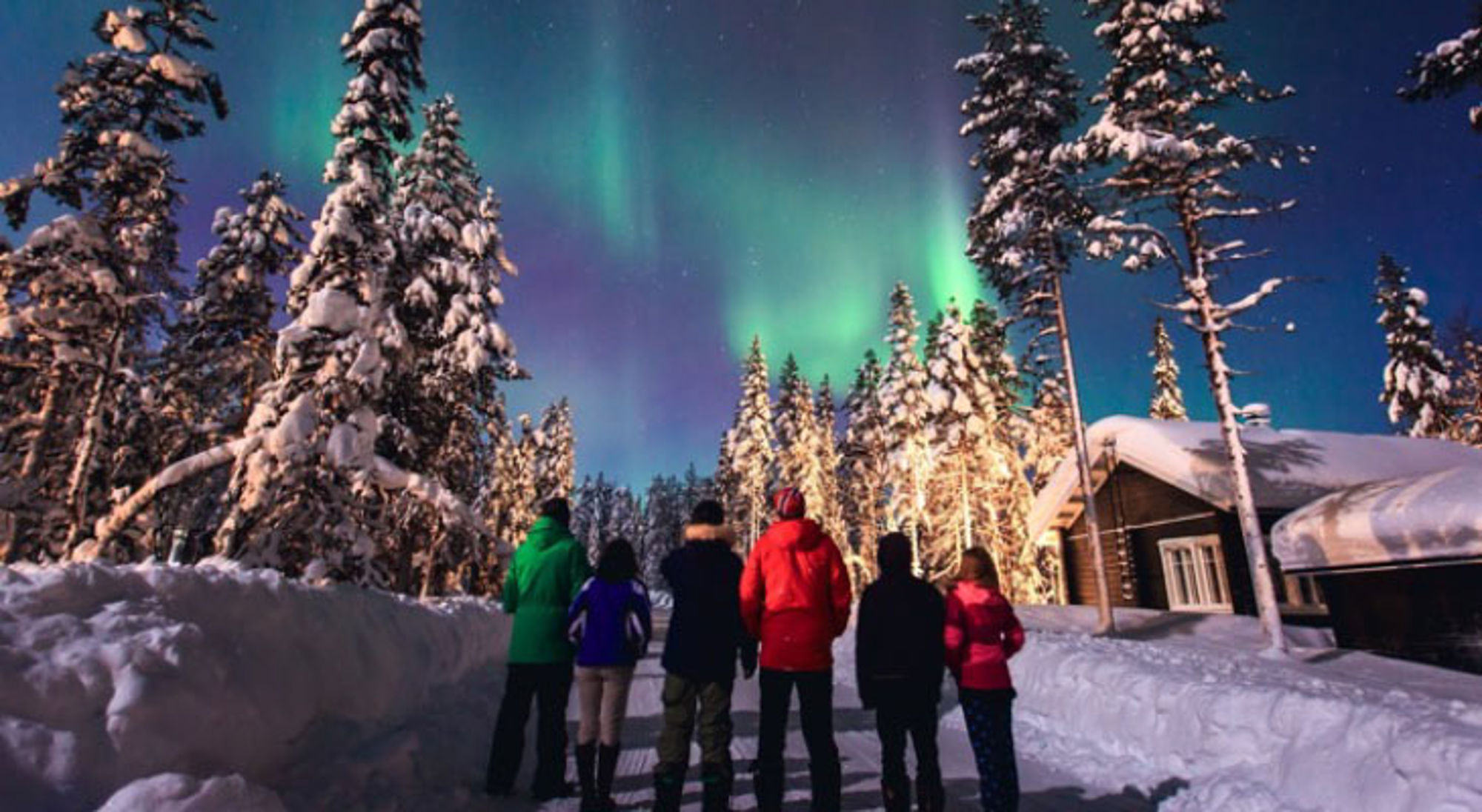 Groep mensen kijkt naar het noorderlicht boven een besneeuwd bos en houten lodge.