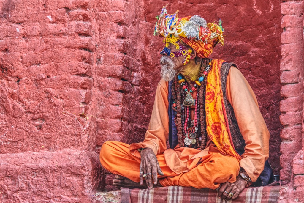 Sadhu in kleurrijke kleding en sieraden zit tegen een rode muur in Nepal.