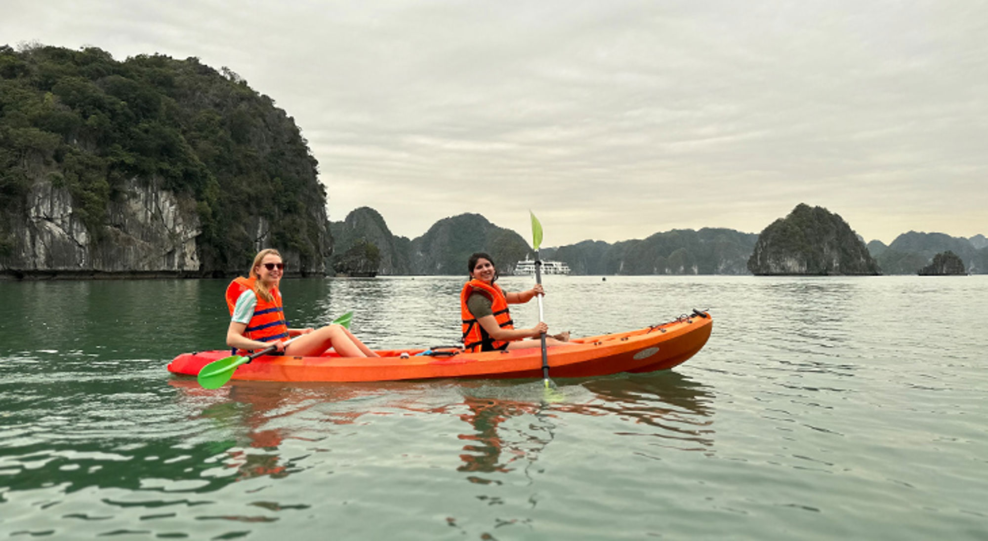 La Han Bay Vietnam Twee reizigers kajakken in een oranje kajak tussen kalkstenen rotsen op rustig water.