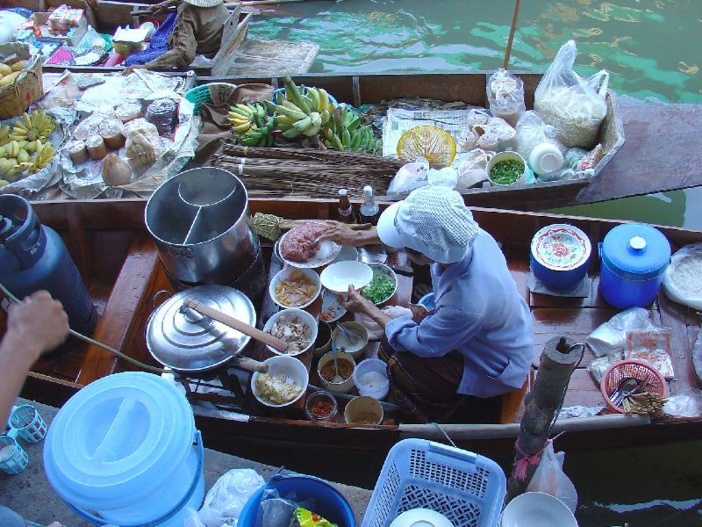 Drijvende markt in Thailand met verkoper die ingrediënten bereidt in een boot tussen fruit en etenswaren.