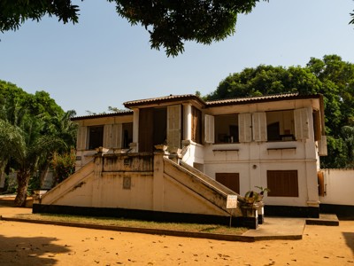 Wit gebouw met buitentrap en houten luiken, omringd door bomen tijdens reisroute Ghana, Togo en Benin dag 7.