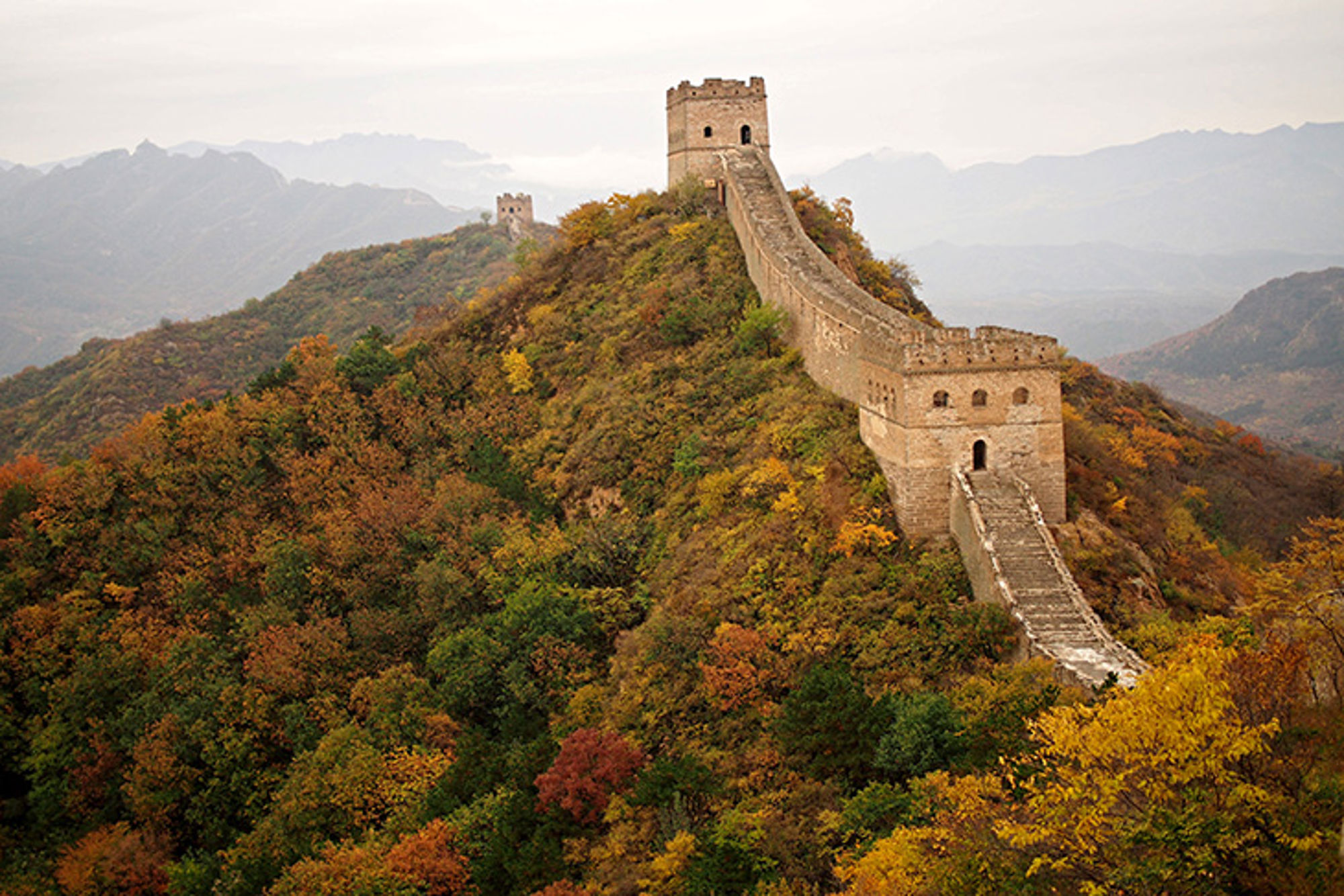 Chinese Muur slingert over beboste heuvels in herfstkleuren, met wachttorens en bergen op de achtergrond.