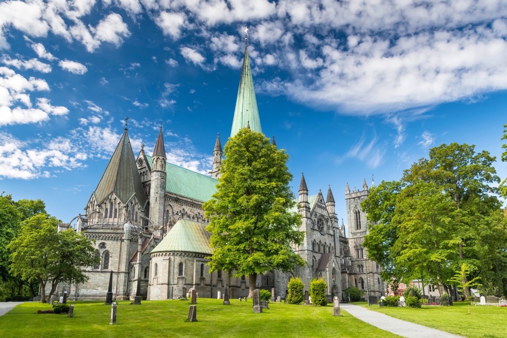 Nidarosdom in Trondheim, Noorwegen, met groene torenspits en park onder blauwe lucht.