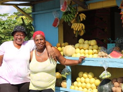 Twee vrouwen bij een fruitkraam met bananen, meloenen en ananas langs de route in Jamaica.