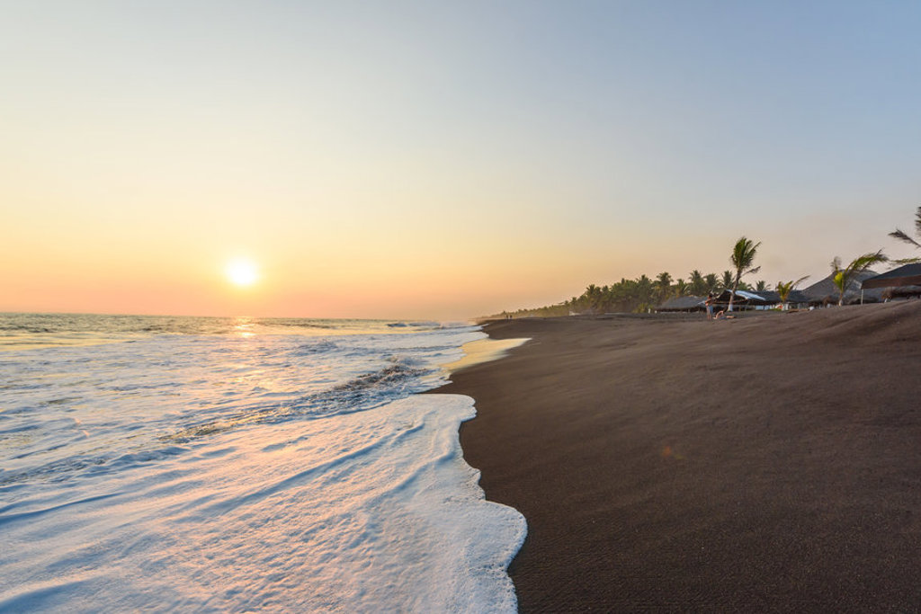 Zonsondergang aan het zwarte zandstrand van Guatemala met palmbomen en golven langs de kust.