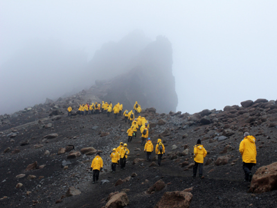 Wandelaars in gele jassen op rotsachtig Antarctisch eiland tijdens Route Antarctica, met mist en kliffen op de achtergrond.