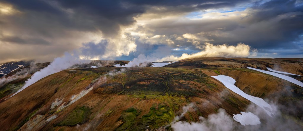 Geothermische stoompluimen boven de heuvels van de Laugavegur Trail in IJsland, met sneeuwvelden en wolkenlucht.