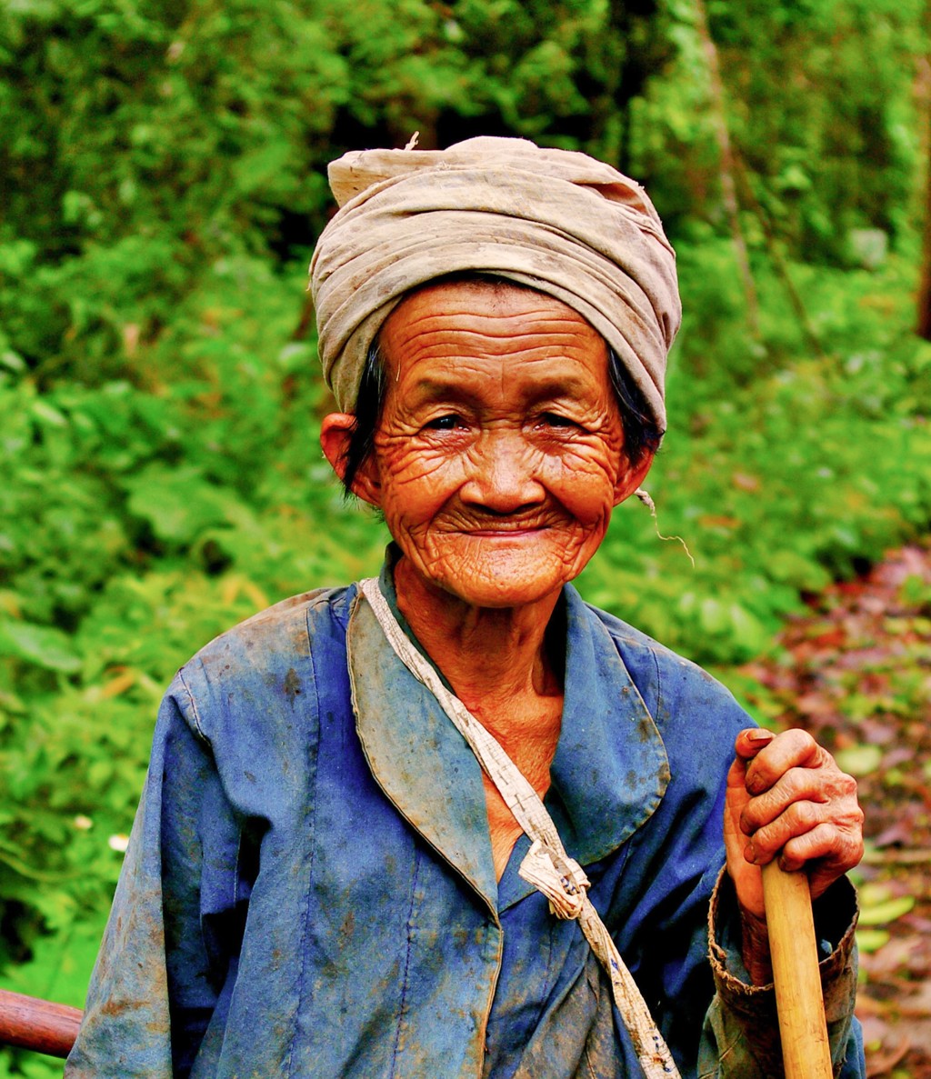 Portret van een glimlachende vrouw met hoofddoek en wandelstok in groen landschap in Thailand.