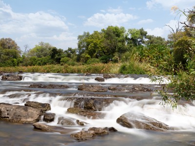 Stromende rivier met kleine watervallen en rotsen, omringd door groen langs de route Victoria Falls - Windhoek.