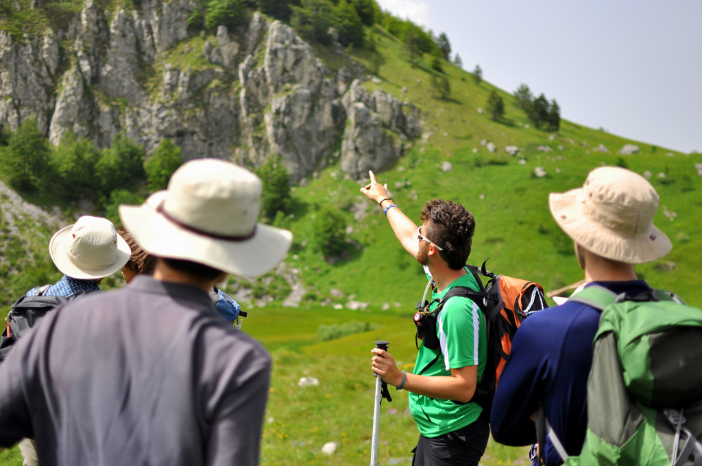 Wandelaars met rugzakken in groene bergen, met rotswanden op de achtergrond tijdens een wandelvakantie.