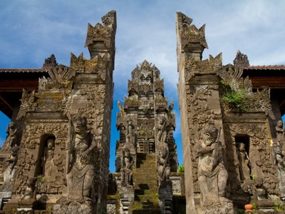 Traditionele Balinese tempelpoort met gedetailleerde stenen reliëfs en beelden onder een blauwe lucht.
