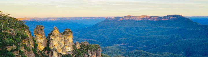 Uitzicht op de Three Sisters rotsformatie in de Blue Mountains, Australië, met groene valleien en kliffen bij zonsondergang.