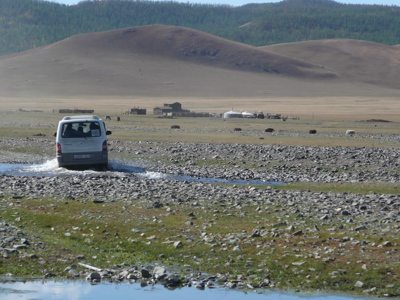 Minibus steekt een ondiepe rivier over in de Mongoolse steppe, met yurts en heuvels op de achtergrond.