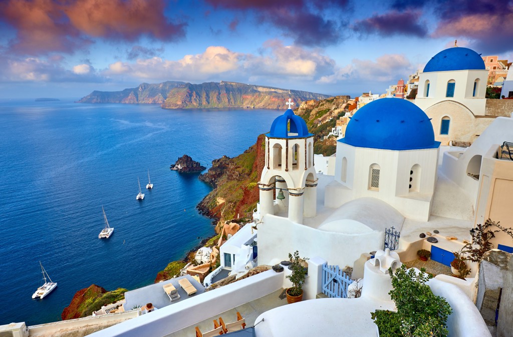 Blauwe koepelkerk en witte huizen in Oia op Santorini met uitzicht op de caldera en zeilboten.