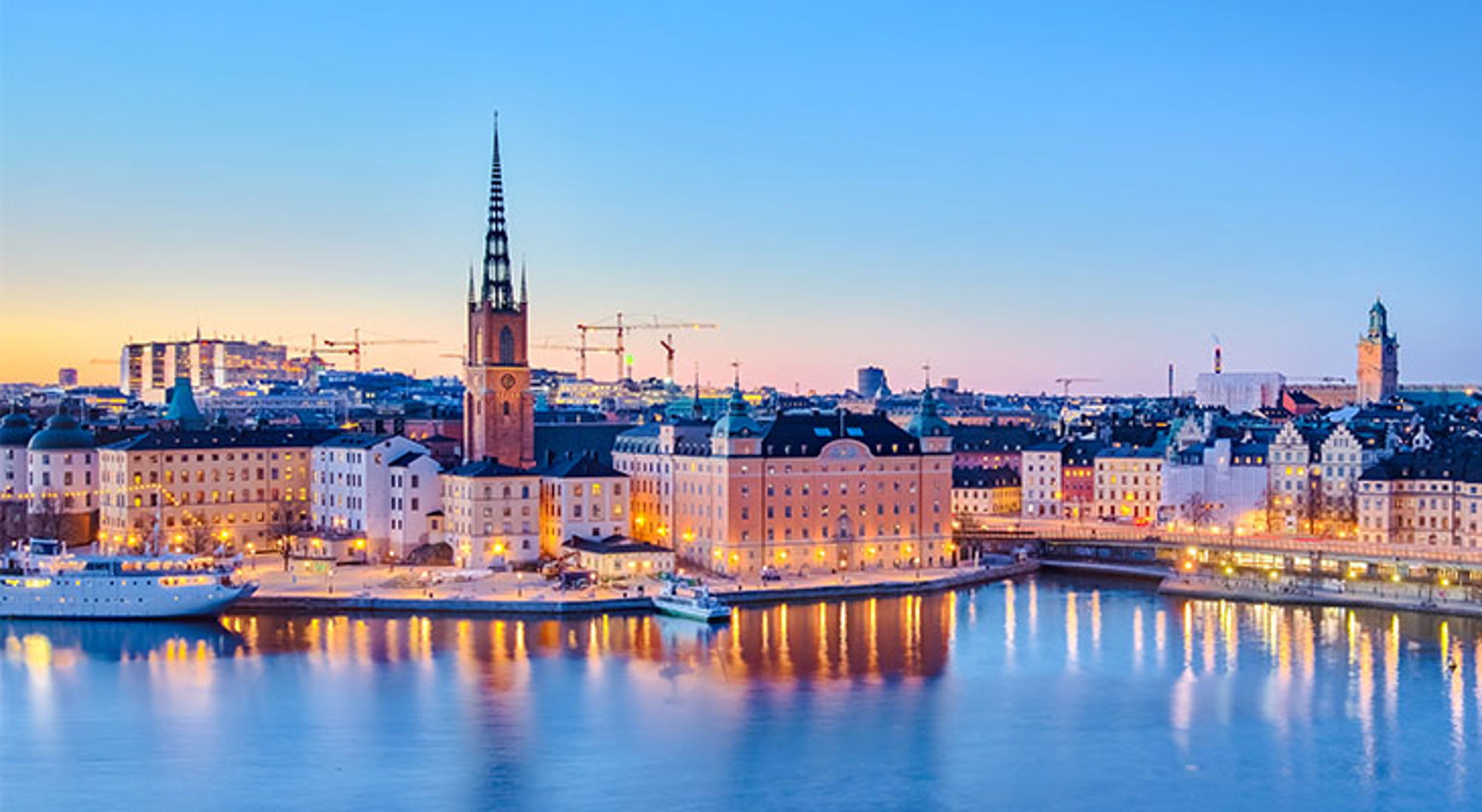 Skyline van Stockholm bij zonsondergang met de Riddarholmskyrkan en verlichte kade langs het water.