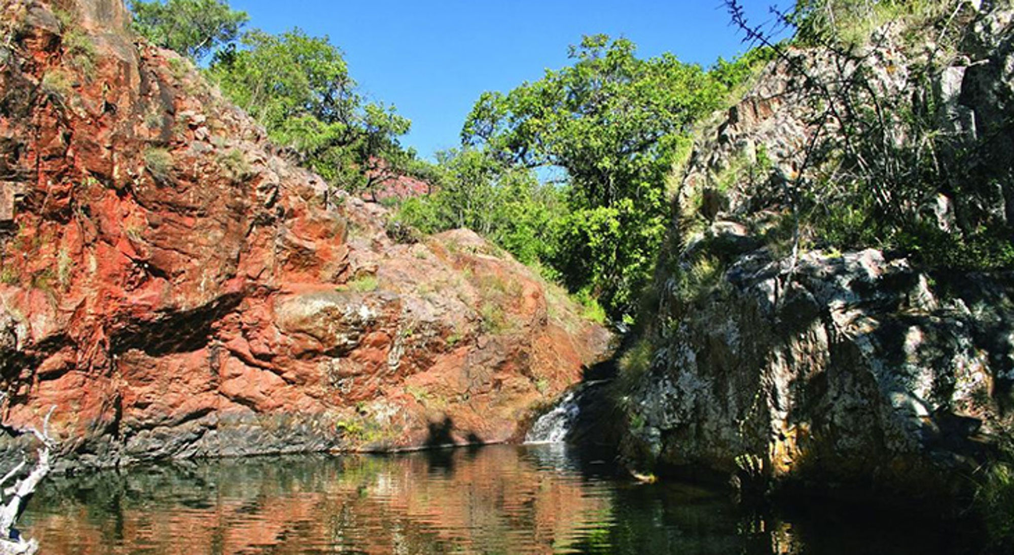 Mashovhela Rock Pool, Zuid-Afrika Rotskloof met rood gesteente, groen struikgewas en een rustig waterbassin onder een blauwe lucht.