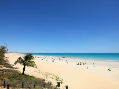 Brede zandstrand aan de kust van West Australië met turquoise zee, parasols en een palmboom op de voorgrond.