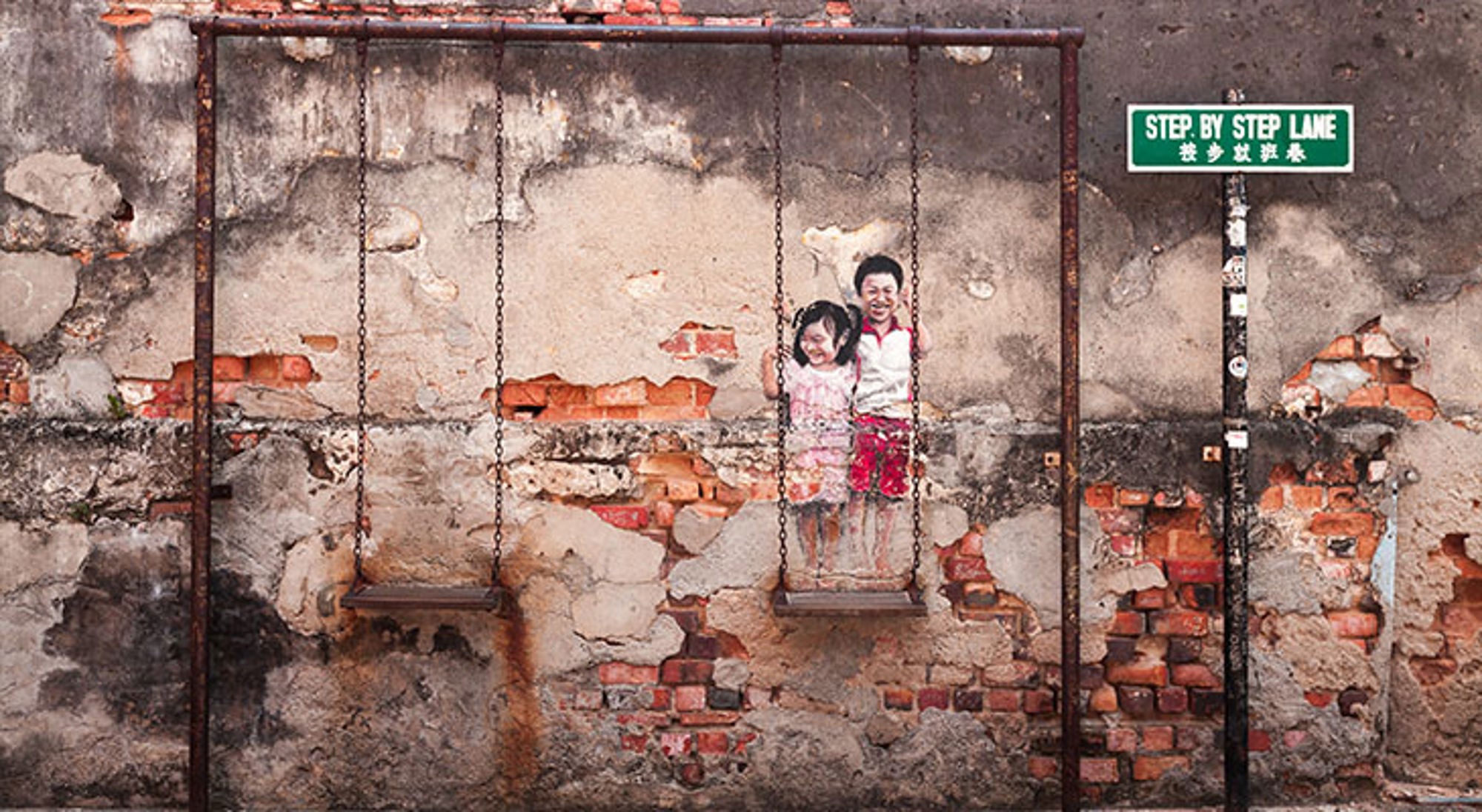 Street art George Town Twee kinderen op een schommel voor een verweerde bakstenen muur, met straatnaambord Step By Step Lane.