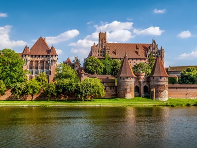 Kasteel Malbork aan de rivier de Nogat in Polen, met rode torens en stadsmuren onder blauwe lucht.