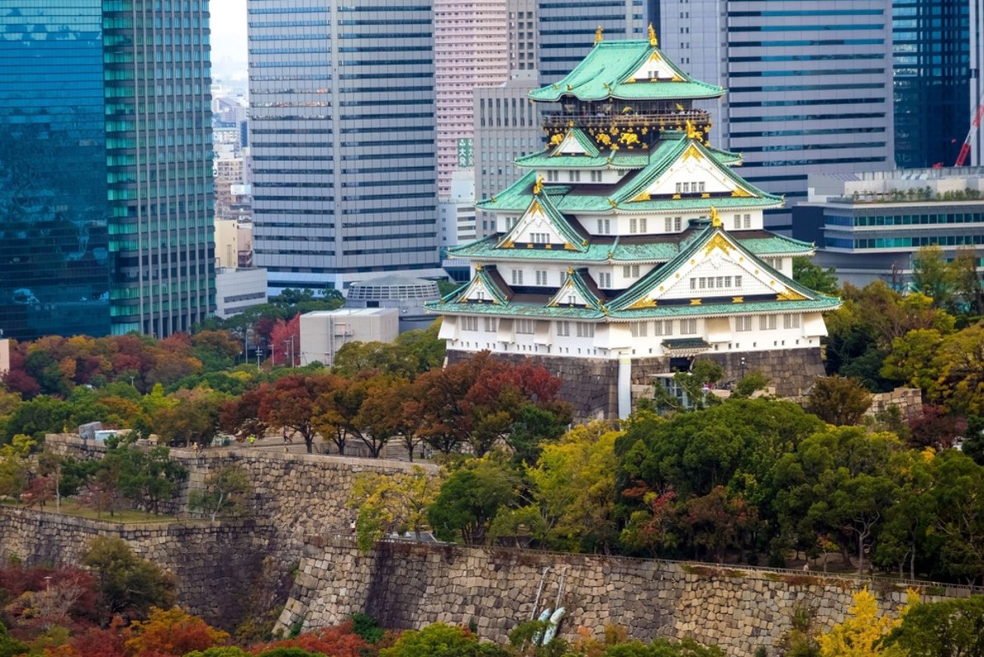 Osaka Castle in Japan met groen dak, omringd door herfstkleuren en moderne wolkenkrabbers.