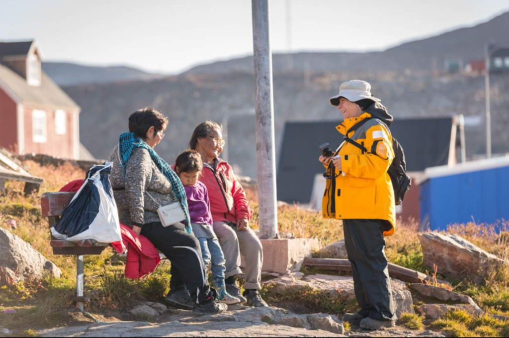 Reiziger in gele jas praat met locals op een bankje in een Groenlands dorp tijdens groepsrondreis.