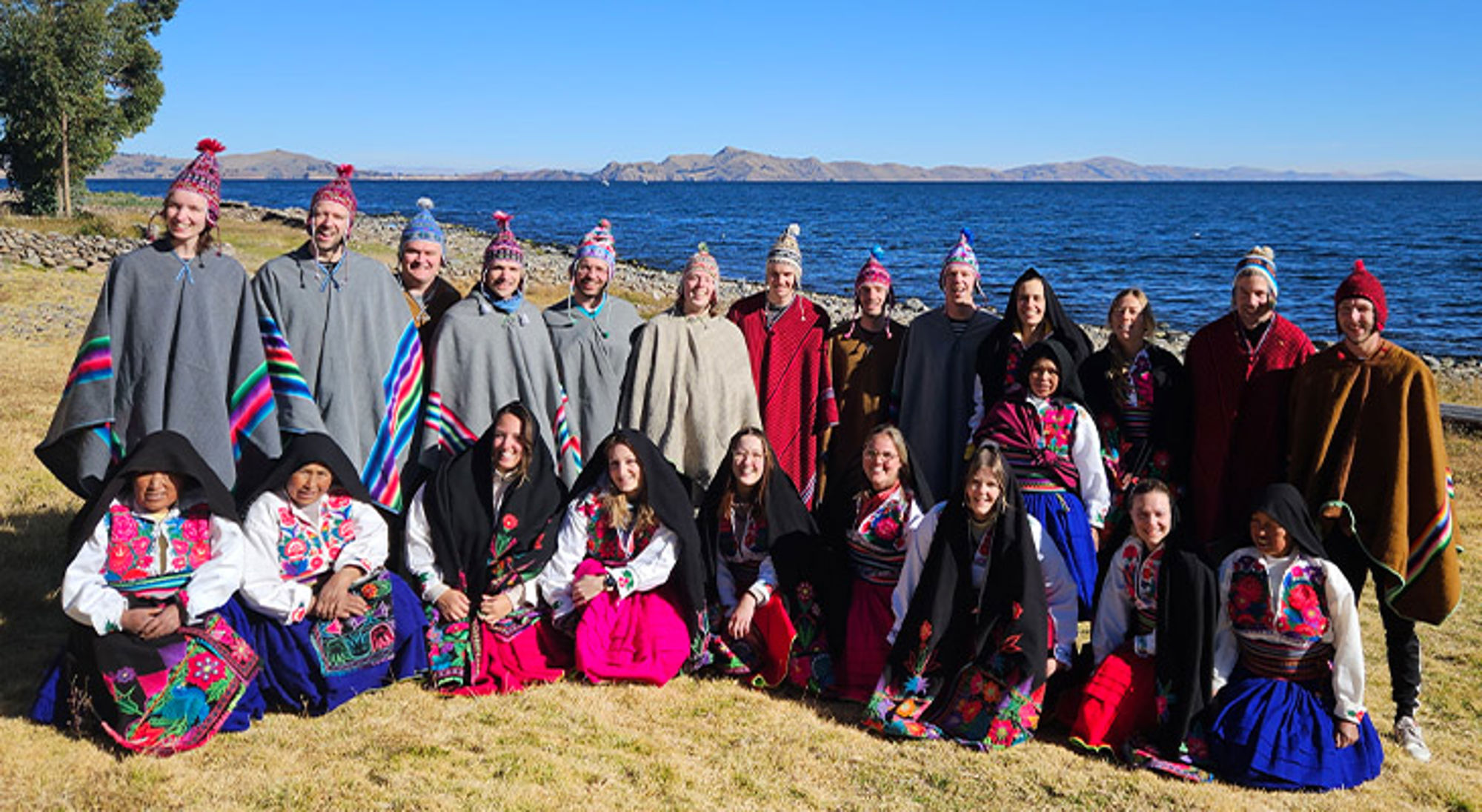 Amantaní Groepsfoto aan het Titicacameer met mensen in kleurrijke traditionele kleding en poncho’s, met bergen op de achtergrond.