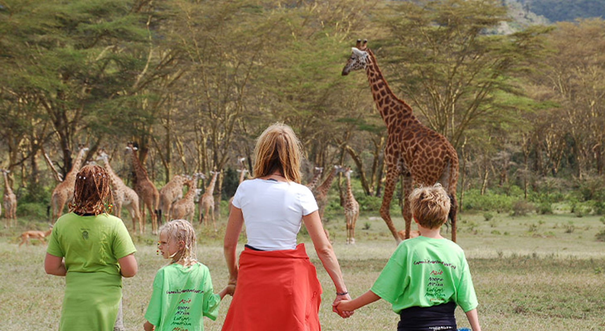Familiereis Kenia Gezin wandelt door savanne met giraffen op de achtergrond tijdens een safari.