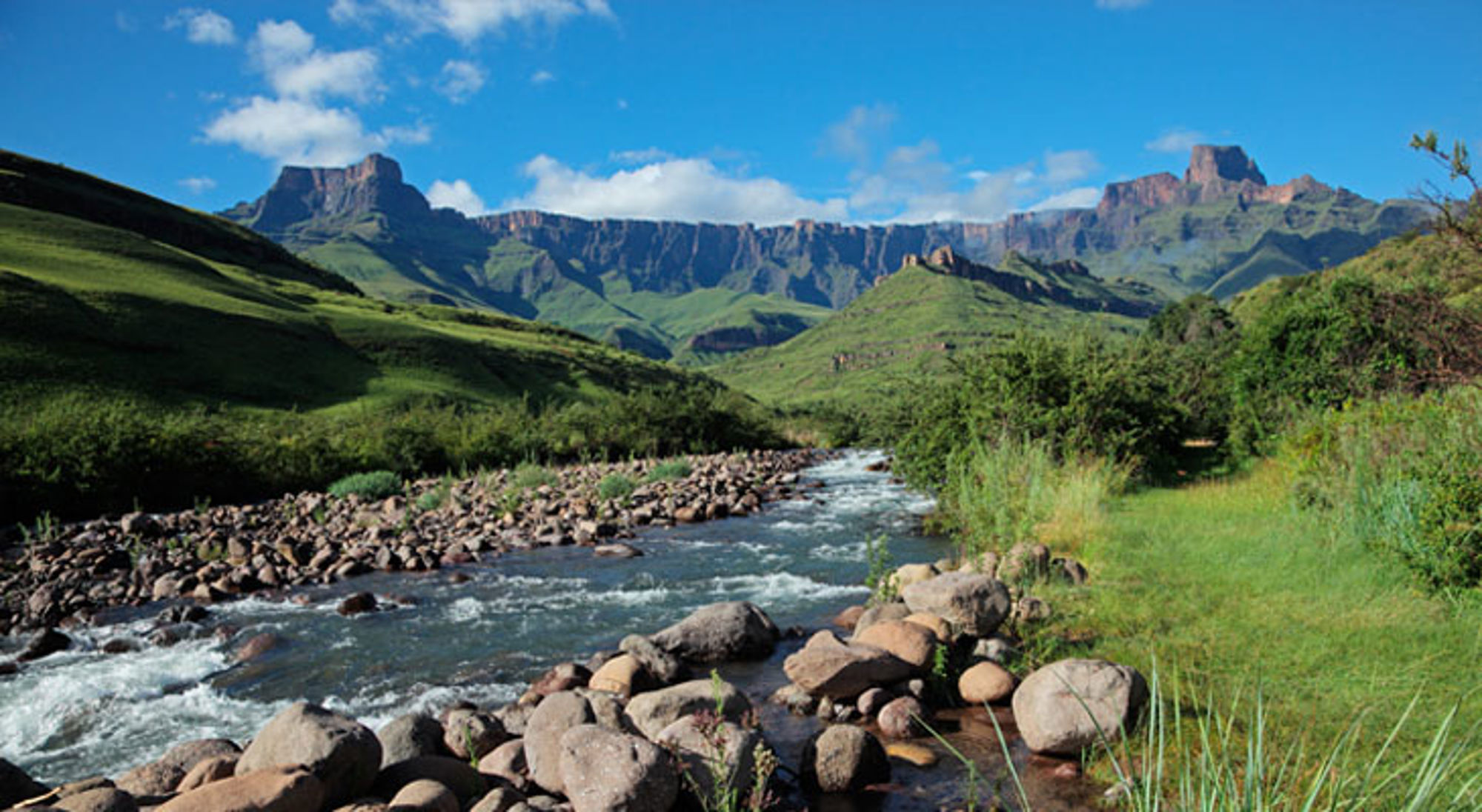Groene Drakensbergen in Zuid-Afrika met een snelstromende rivier en rotsen onder een helderblauwe lucht.