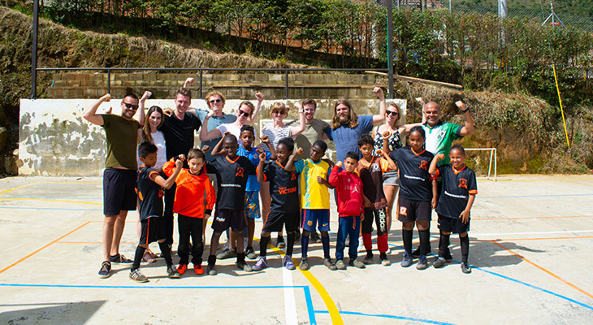 Doneer aan lokale projecten met een positieve impact Groepsfoto van kinderen en volwassenen op een sportveld, met opgestoken armen na een voetbalactiviteit.