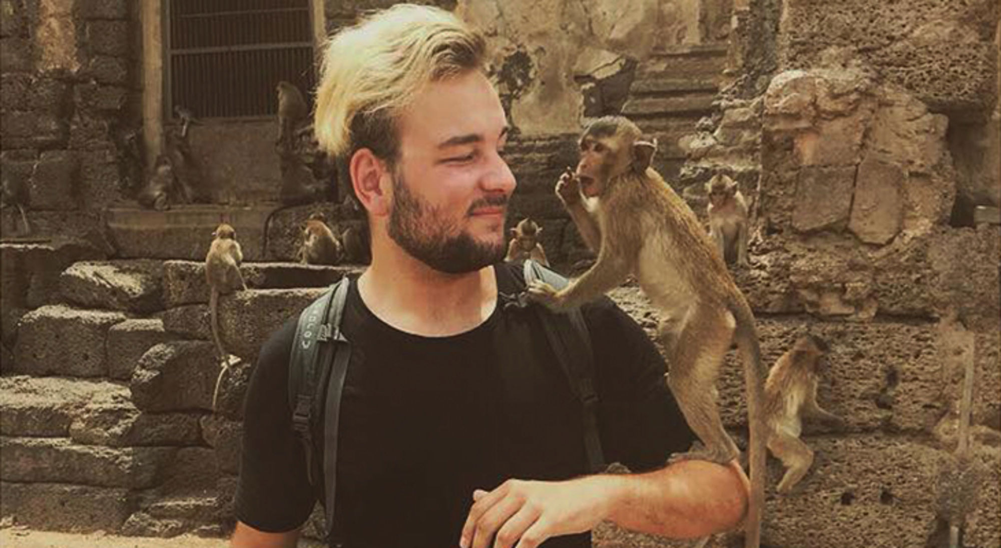 Reiziger met rugzak krijgt bezoek van een aap op zijn arm bij oude tempelruïnes.