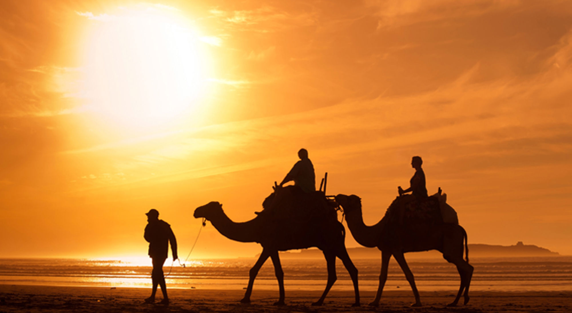 Silhouetten van twee kamelen met ruiters en een gids op het strand bij zonsondergang.