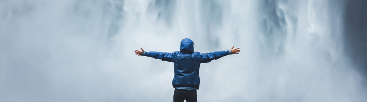 Persoon met gespreide armen voor een enorme waterval met opspattende nevel.