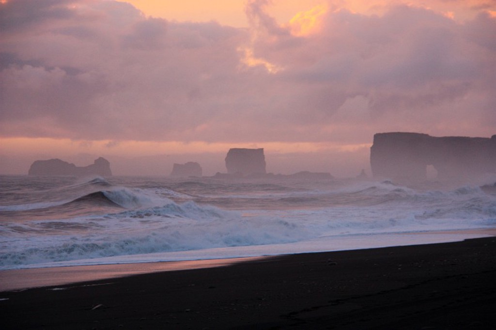 Zwarte zandstrand in IJsland met ruige golven en basaltrotsen bij zonsondergang.