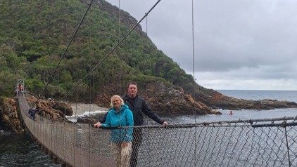 Reizigers op hangbrug langs de kust in Zuid-Afrika, met rotsen en groene heuvels op de achtergrond.