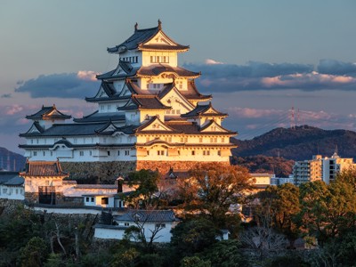 Himeji-kasteel in Japan, verlicht door de ondergaande zon, met heuvels en stad op de achtergrond.
