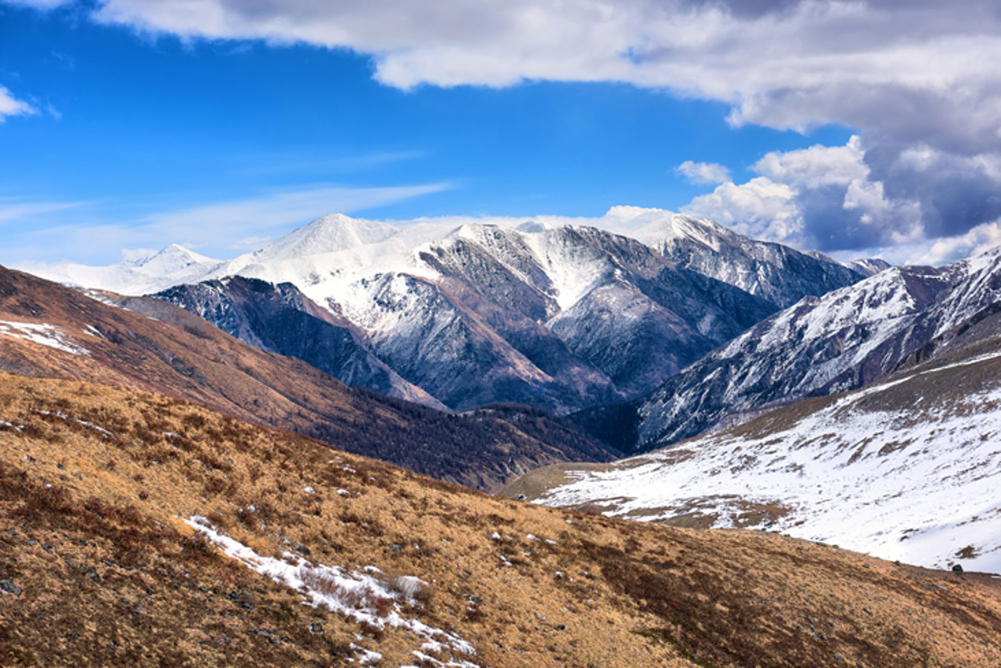 Besneeuwde bergketen met dal en bruine hellingen onder een helderblauwe lucht.