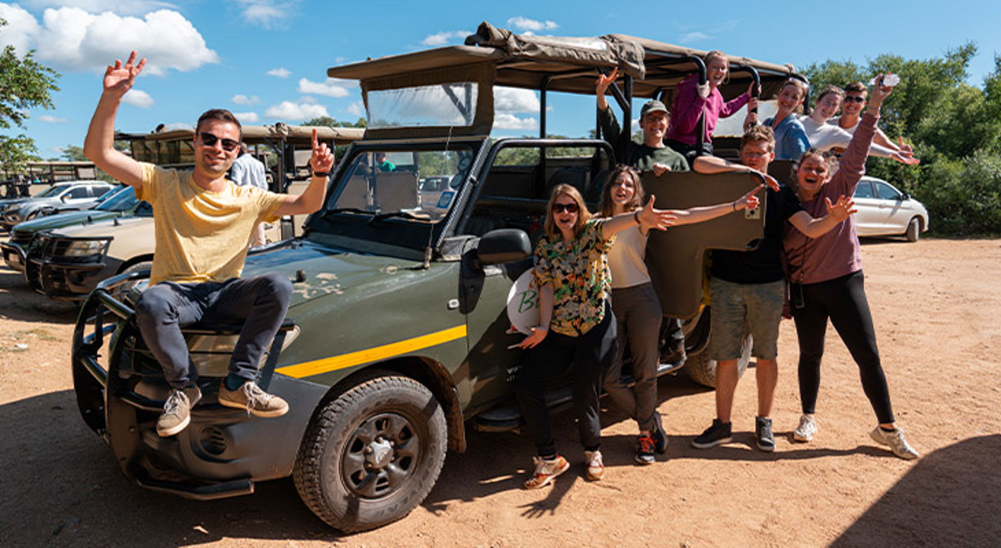 22-35ers groep op safari Groep reizigers zwaait bij een safarijeep op een zonnige dag.