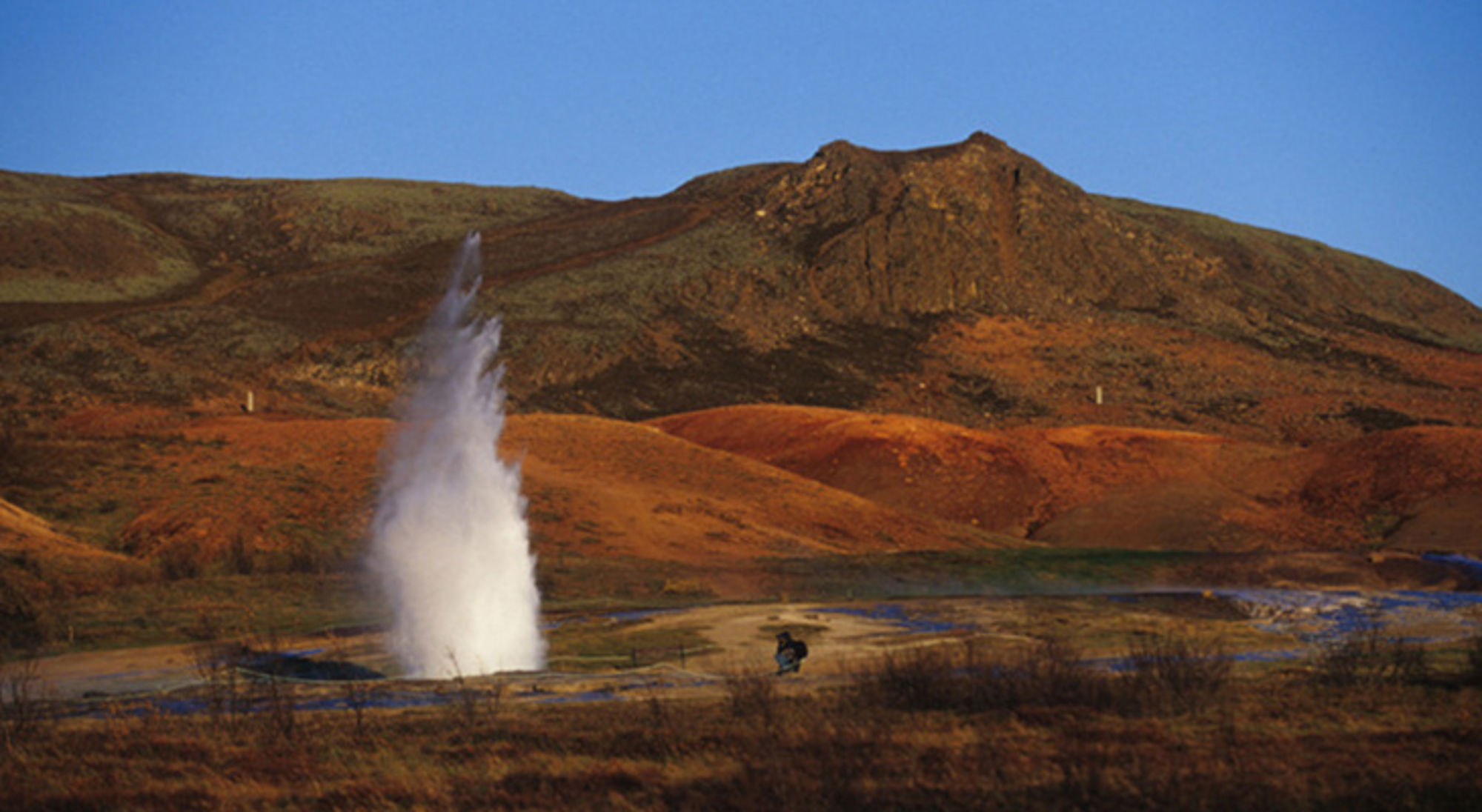Geyser die water en stoom omhoog spuit in een vulkanisch landschap met heuvels onder een heldere blauwe lucht.